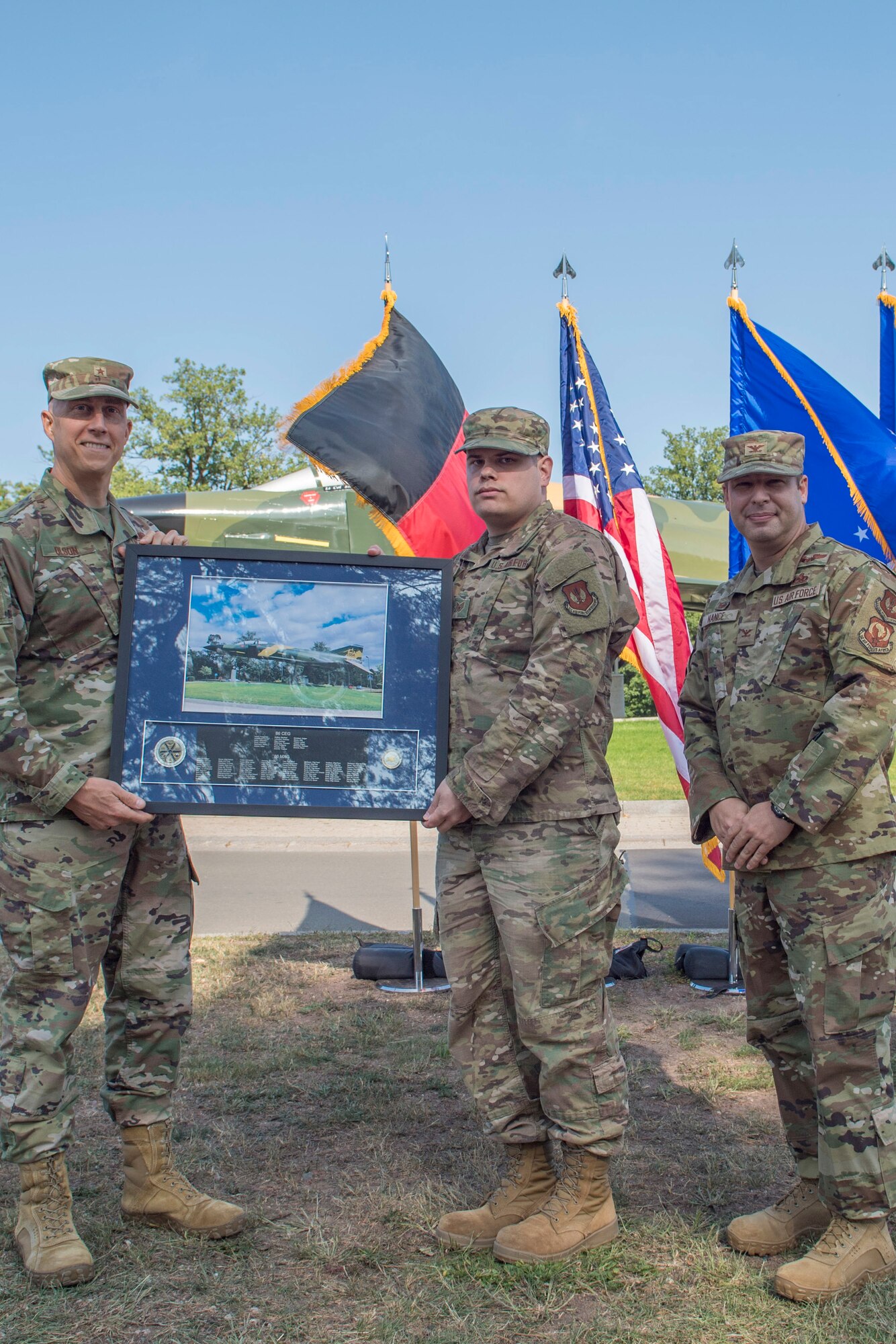 Three Airmen stand in front of an aircraft static display, holding a plaque.