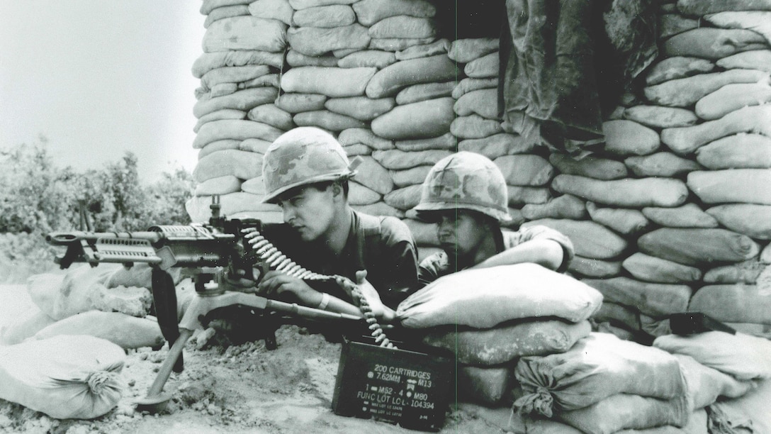 Two service members man a machine gun while surrounded by sandbags.