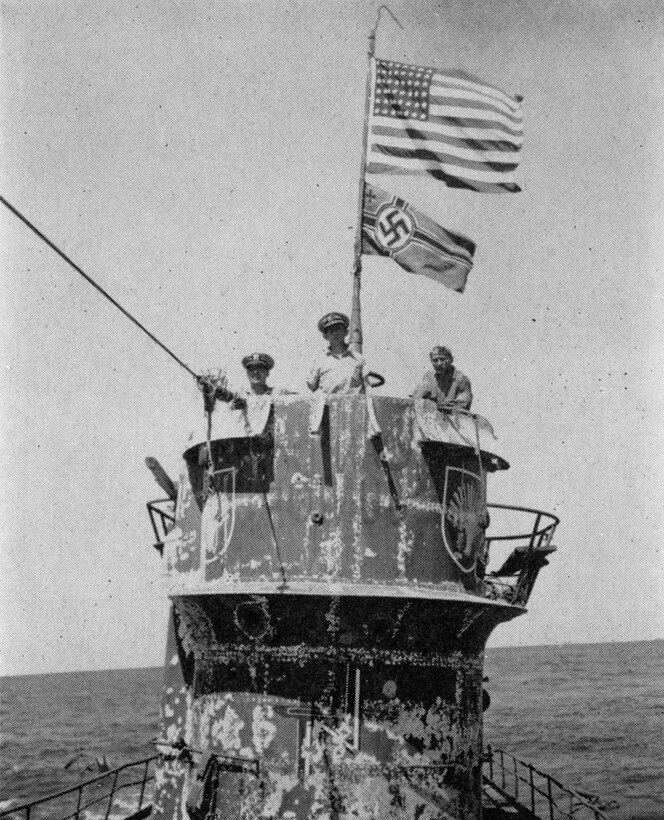 Two flags fly above three men standing on the conning tower of a submarine in the ocean.
