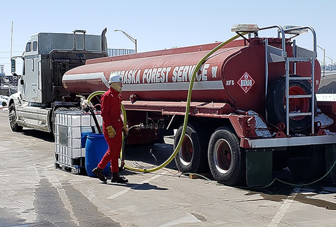 A man in safety equipment unloads a red tanker truck into a cubical container attached to a pallet.