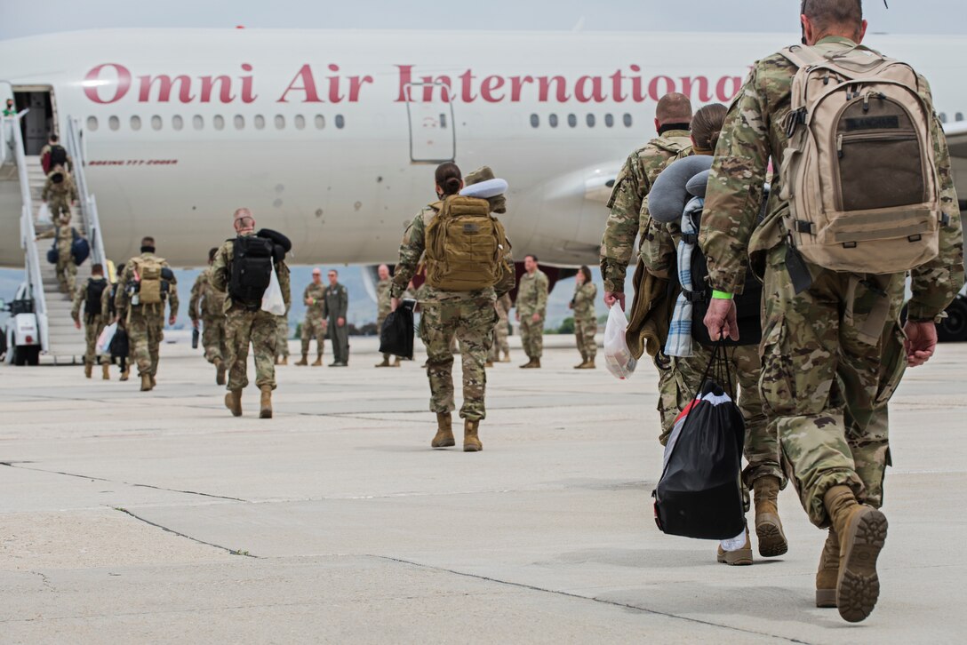 The image is looking at the back of line of Air Force Airmen who are carrying personal items as they walk towards an aircraft staircase to load on a white airliner aircraft with the company name, Omni Air International, written in red letters on the side of the aircraft.