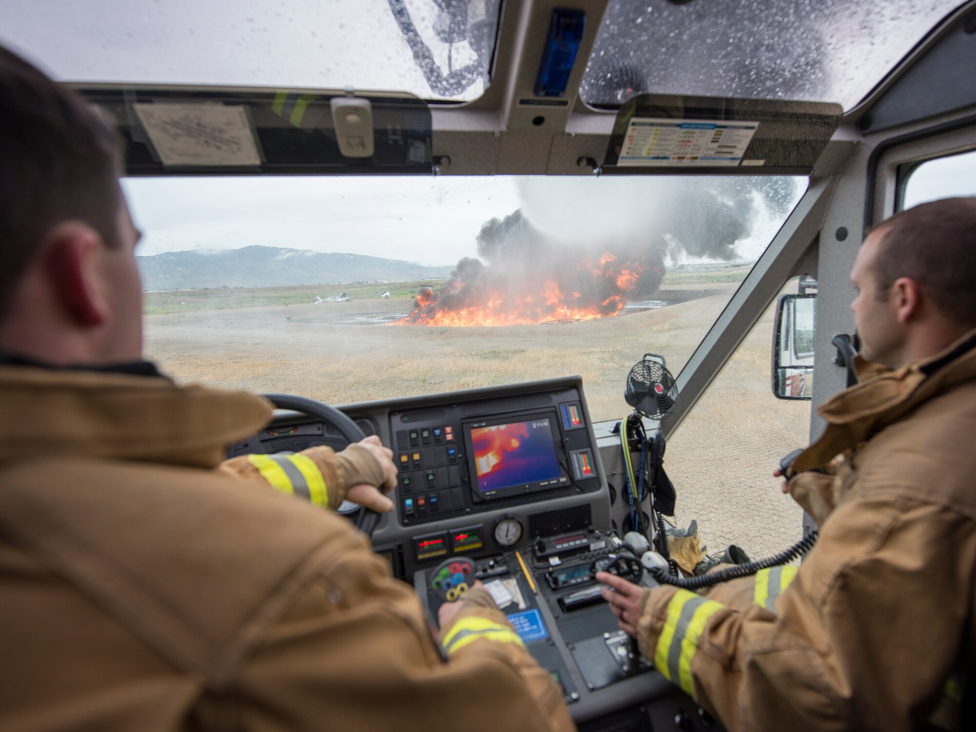 Two male fire fighters in their brown and yellow fire suits sit inside a fire truck that is overlooking a fire with billowing black smoke. Inside the cab of the fire truck is an infrared screen showing the heat from the fire.