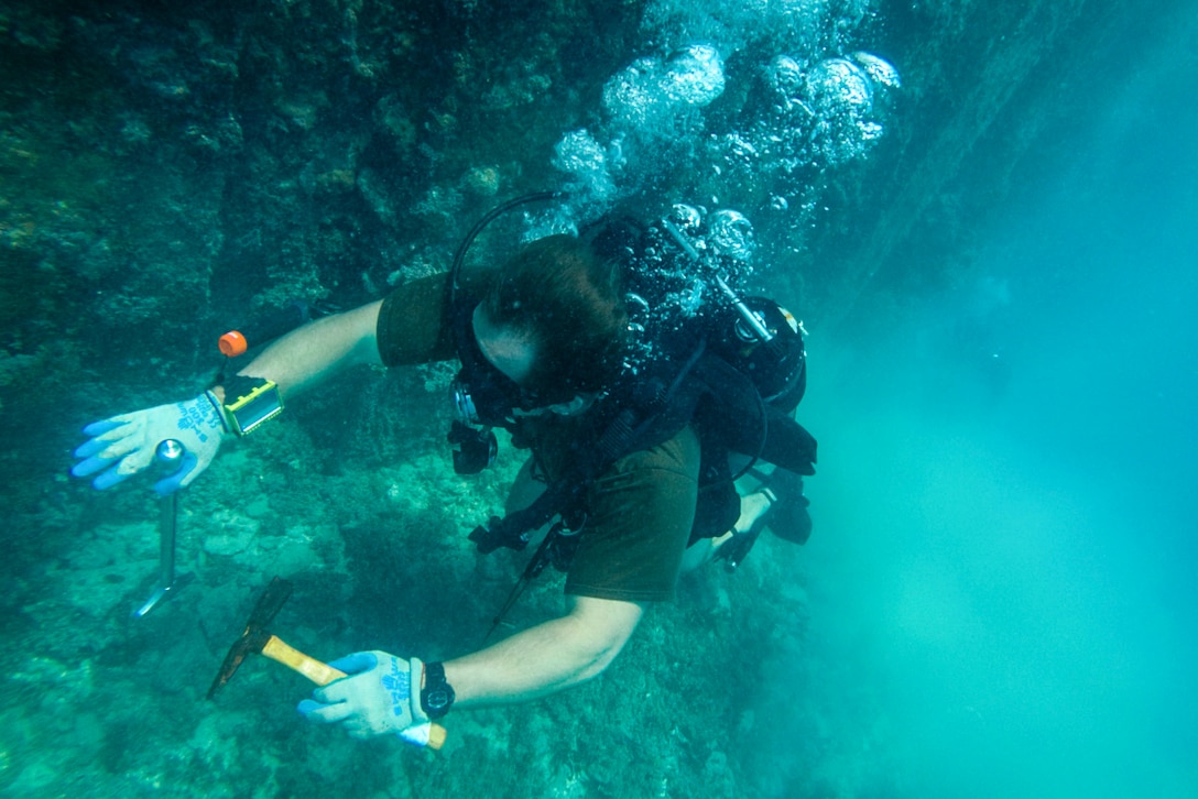 A sailor in dive gear holds tools while working on a structure underwater.