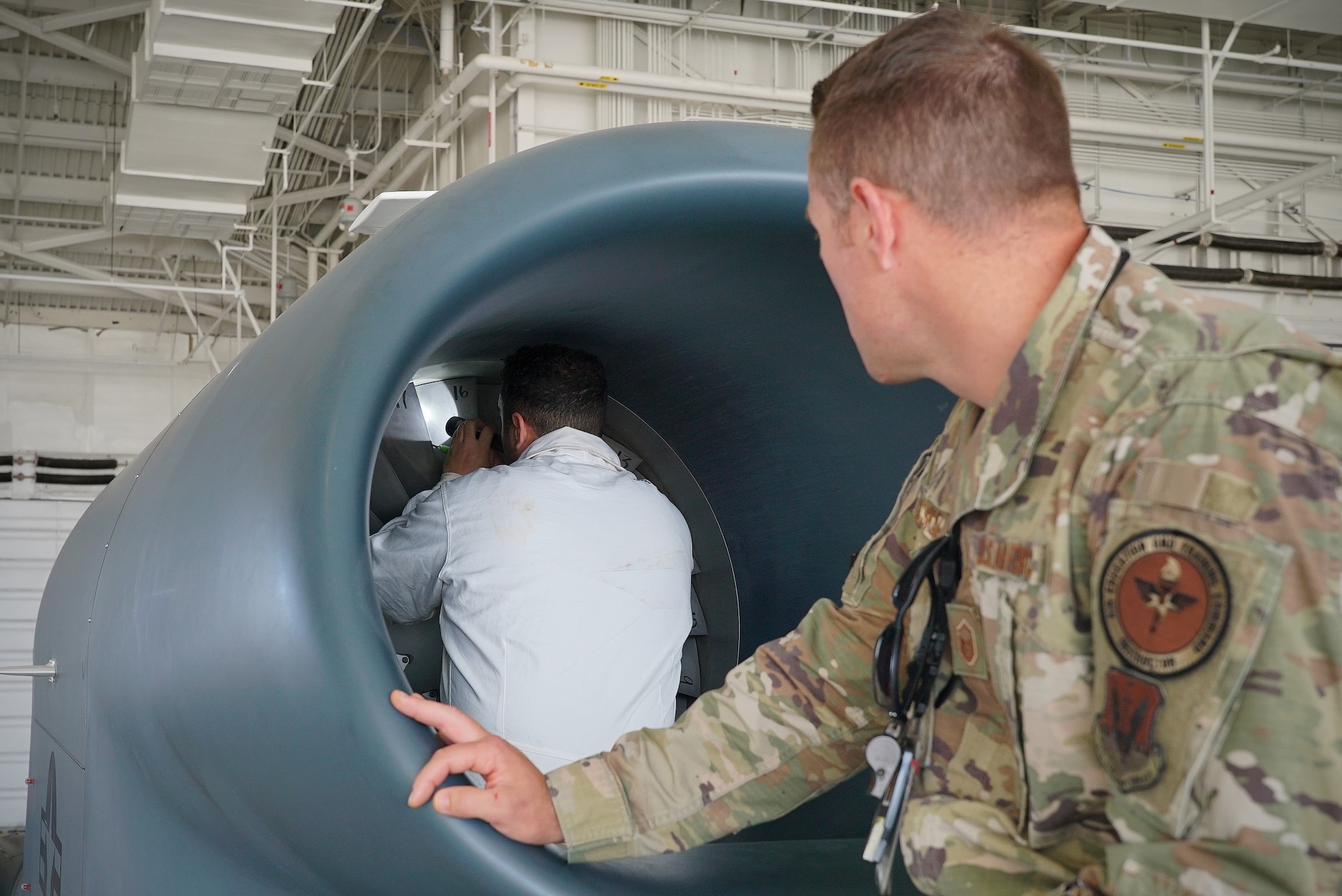 A uniformed military member on the right-hand side of the frame looks on as his coworker in a protective smock inspects the turbine of an aircraft with a flashlight.