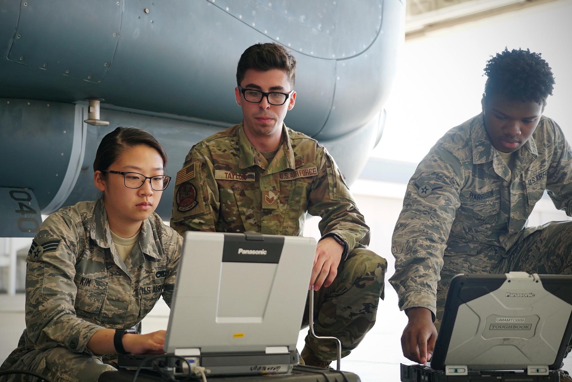 Three uniformed military members kneel down shoulder-to-shoulder in front of portable computers they have placed on the ground.