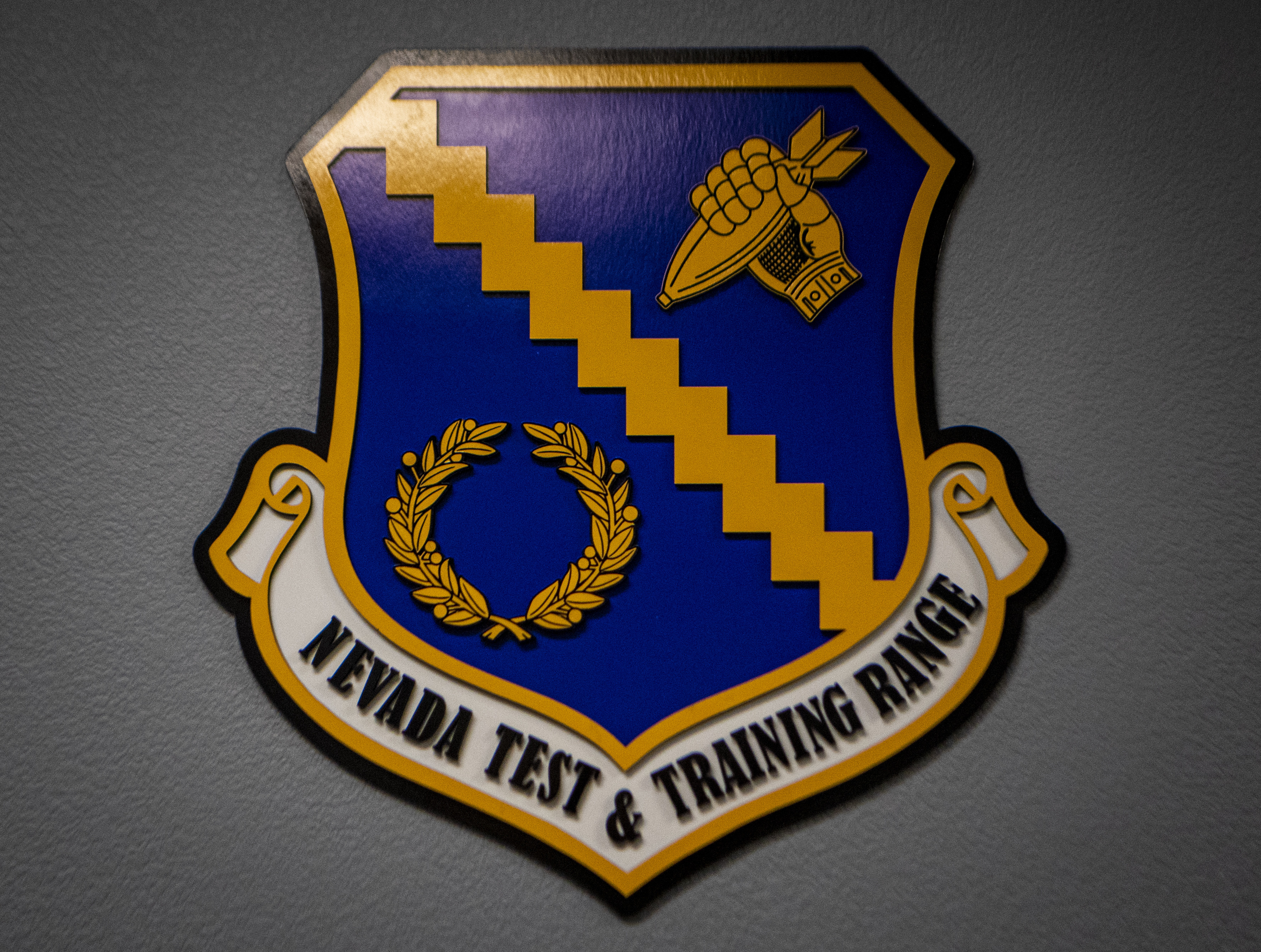 A crest of the Nevada Test and Training Range (NTTR) is displayed in a hallway at the NTTR headquarters building at Nellis Air Force Base, Nevada, Sept. 23, 2020. The NTTR provides warfighters a flexible, realistic and multi-dimensional battlespace to conduct testing tactics development, and advanced training in support of U.S. national interests. (U.S. Air Force photo by Airman 1st Class Dwane R. Young)