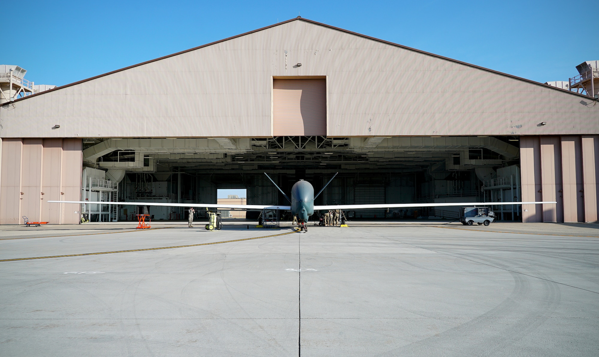 A small unmanned aircraft is shown in center frame in front o f an open hangar, with its wingspan shown in entirety from left to right of the picture.