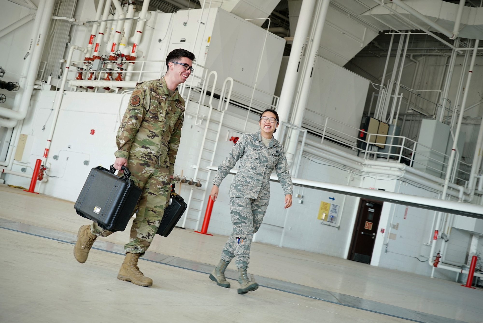 A military-uniformed man and woman walk through a large aircraft hangar next to each other as they smile in conversation.