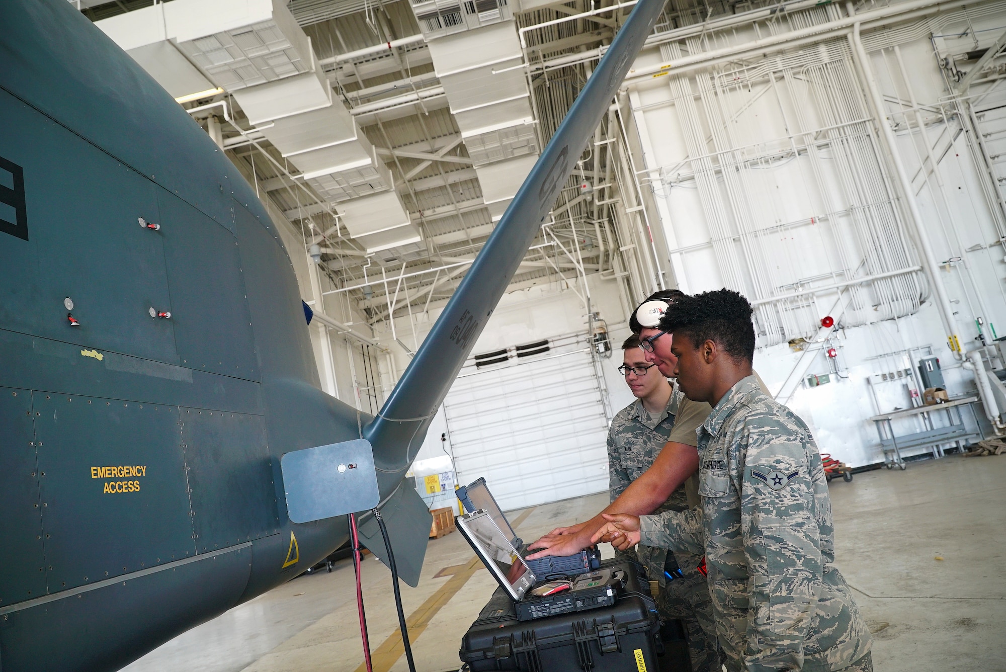 The side of a small unmanned aircraft is shown on the left-hand side of the frame, as three uniformed military members stand near it with a laptop in front of them.