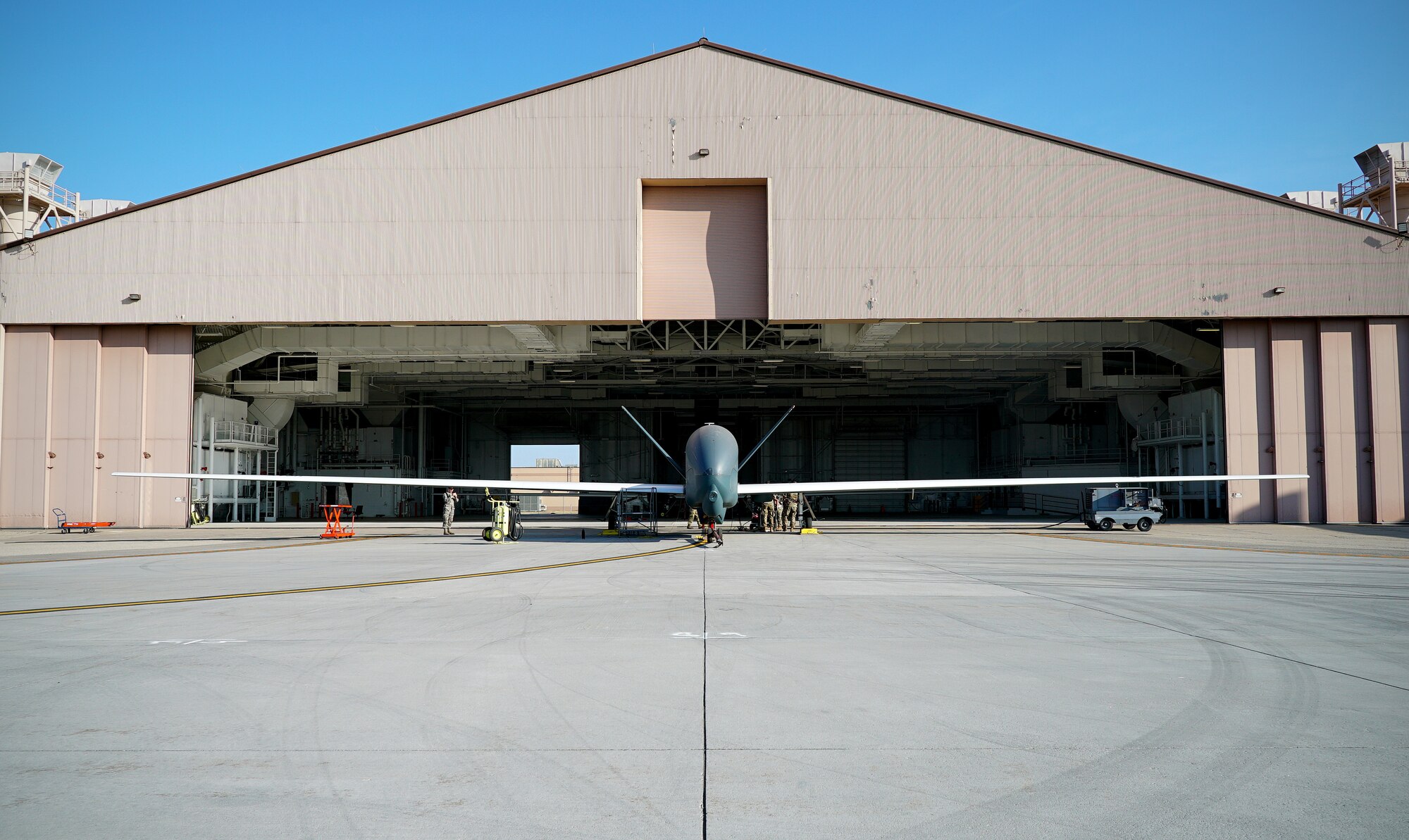 A small unmanned aircraft is shown in center frame in front o f an open hangar, with its wingspan shown in entirety from left to right of the picture.