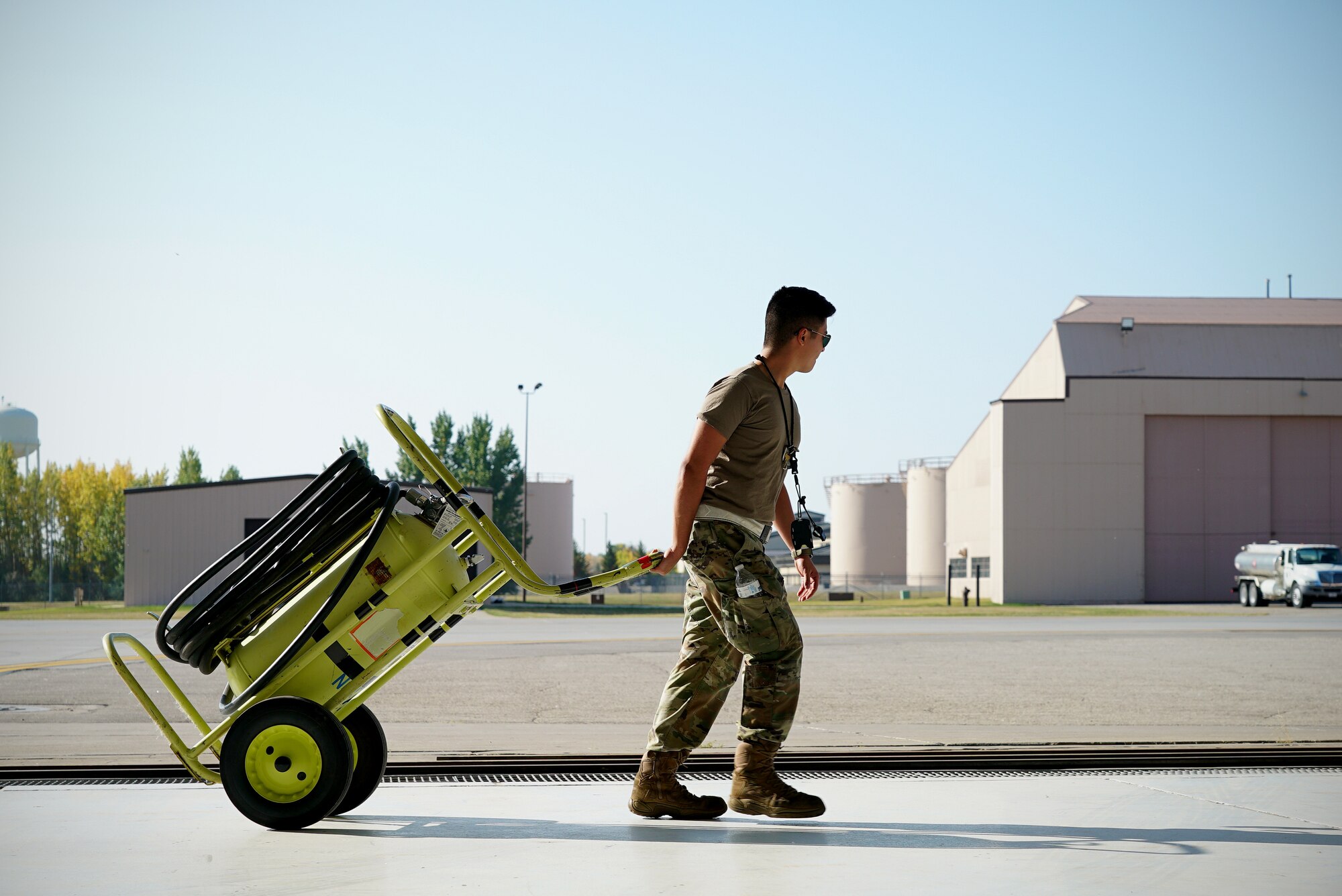 A uniformed military member walks across the frame with a large neon yellow wheeled fire extinguisher in tow.