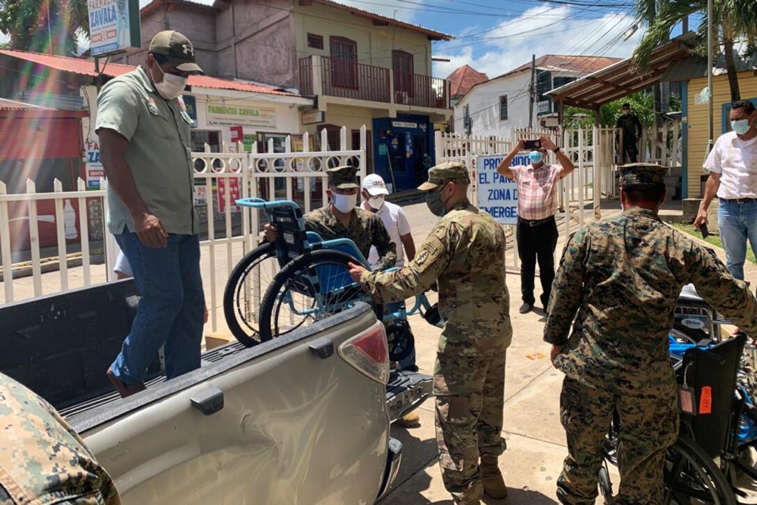 Three service members wearing face masks help unload wheelchairs from the bed of a truck.