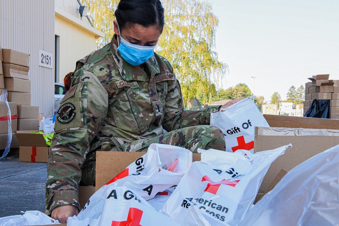 An airman wearing a face mask takes a plastic bag bearing the Red Cross logo out of a box; many other bags with items inside are situated next to the box.