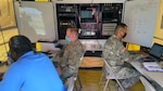 Three men, two in green camo uniforms, sit in a small room with computer equipment.