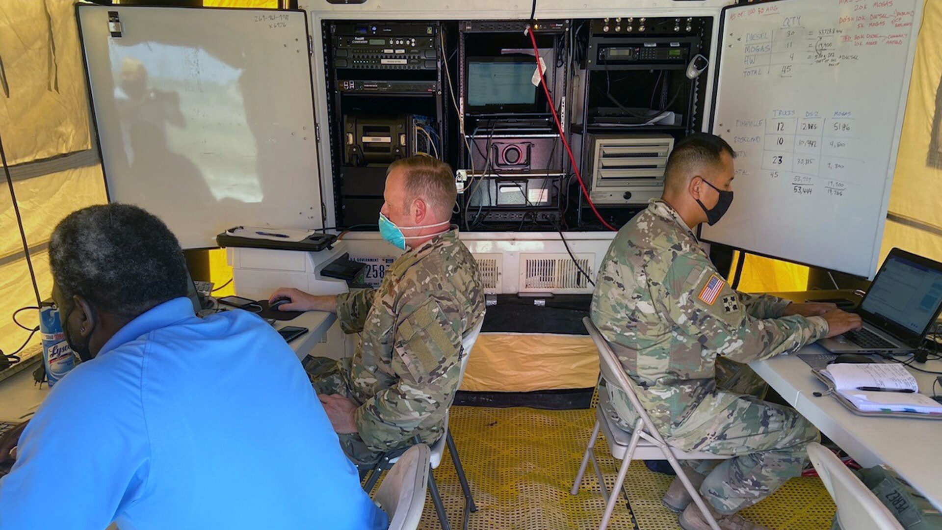 Three men, two in green camo uniforms, sit in a small room with computer equipment.