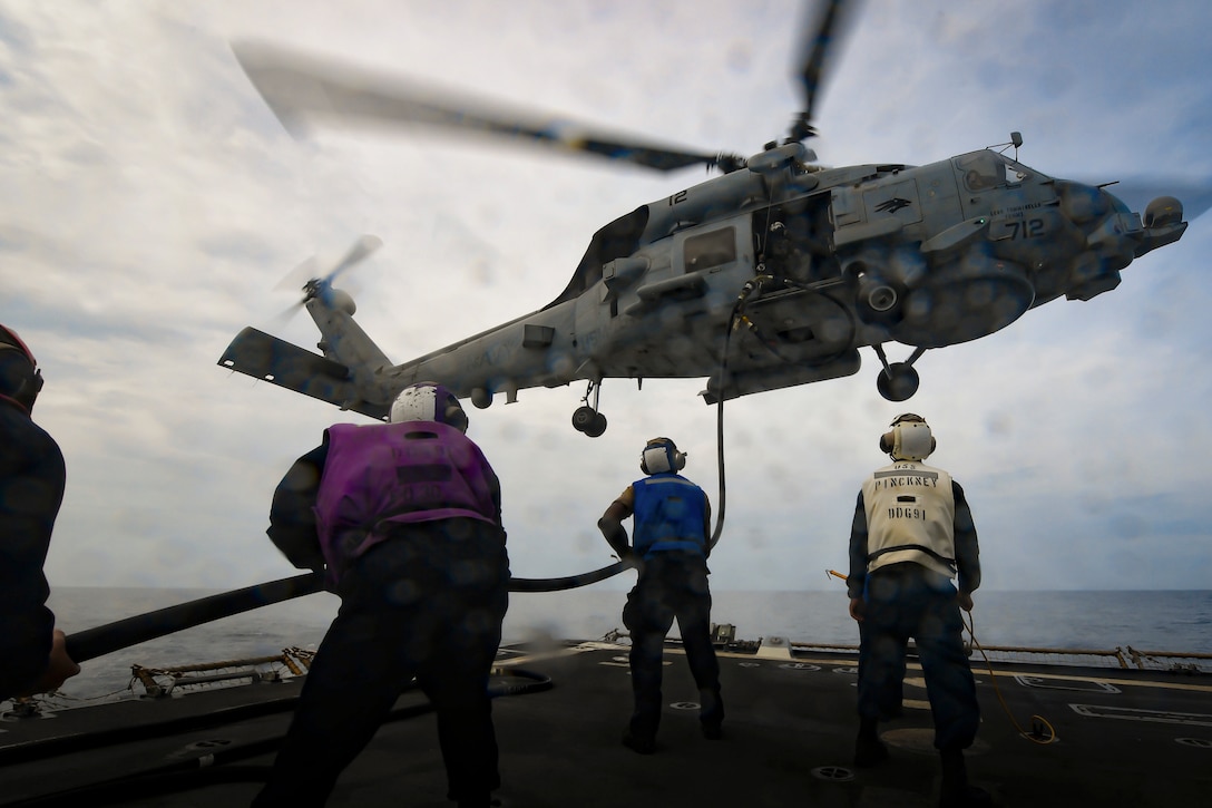 A helicopter hovers as sailors hold a rope aboard a ship.