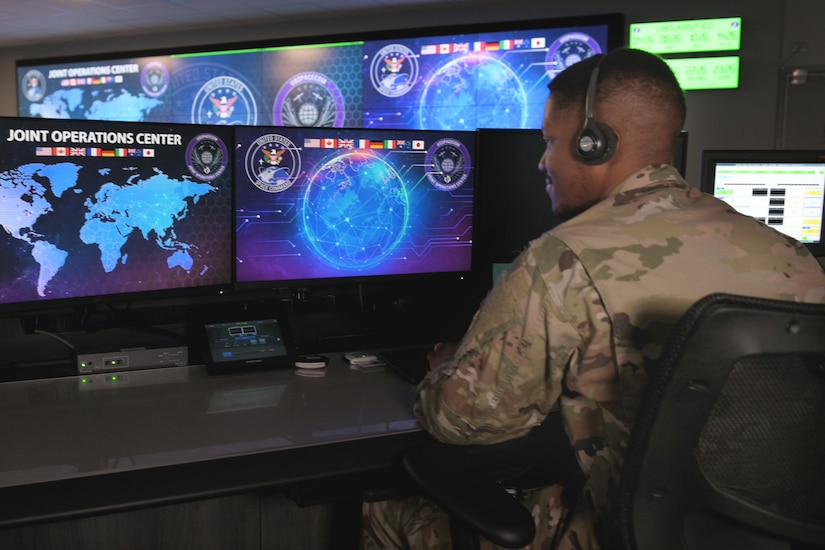 A Space Command service member sits and looks at monitors in a room.