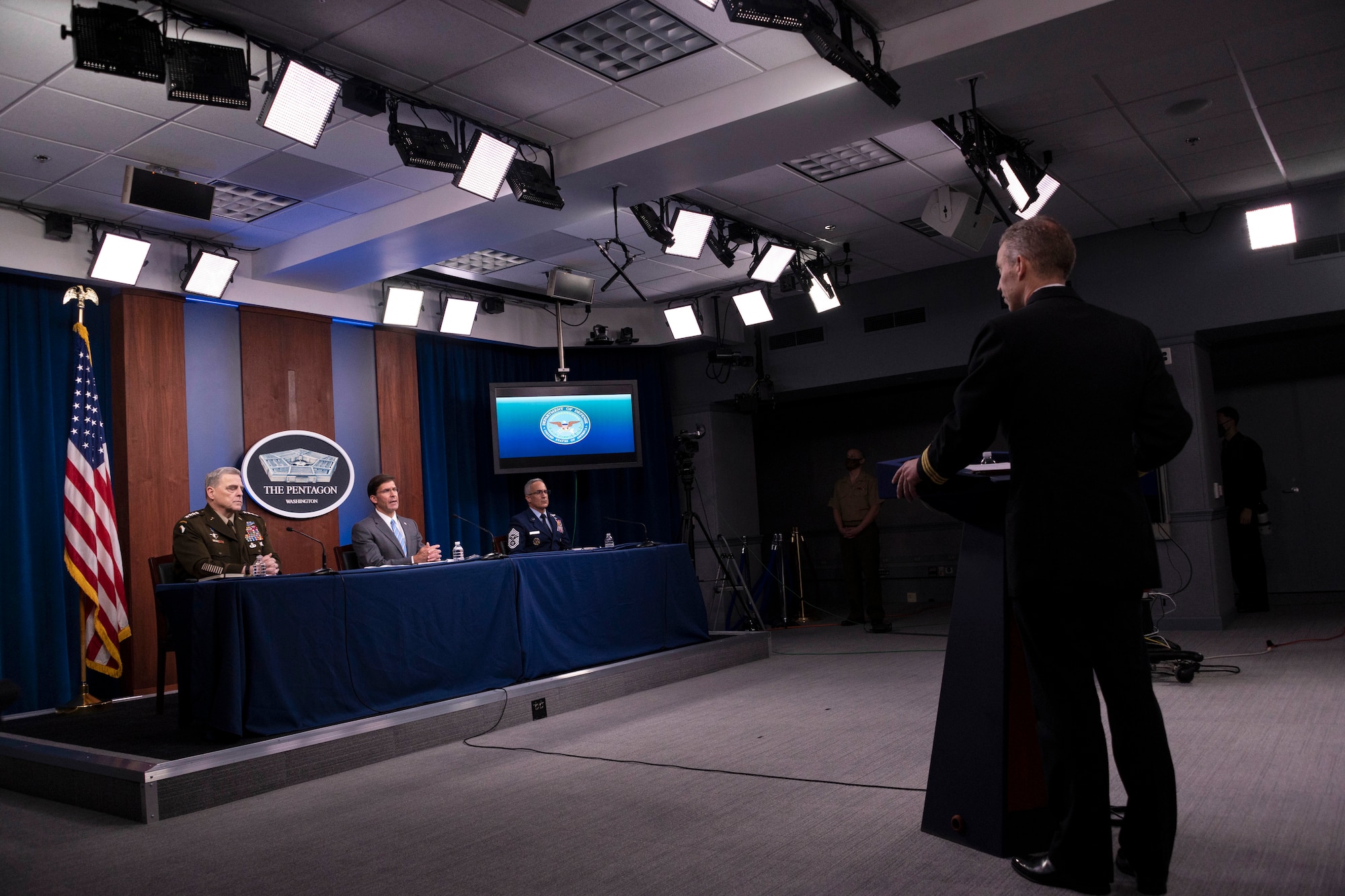 Three Defense Department leaders sit at a table and another person stands at a lectern  in a briefing room.
