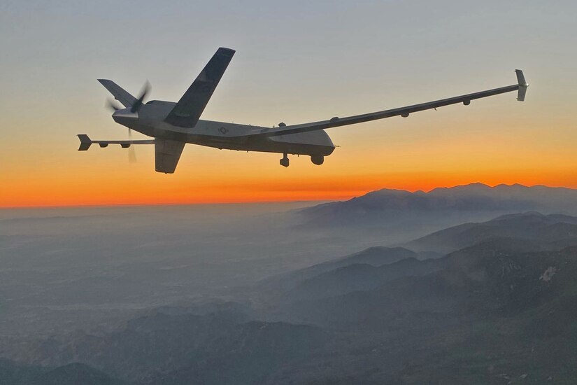 A small military plane flies above a smoky mountain range as the sun sets.