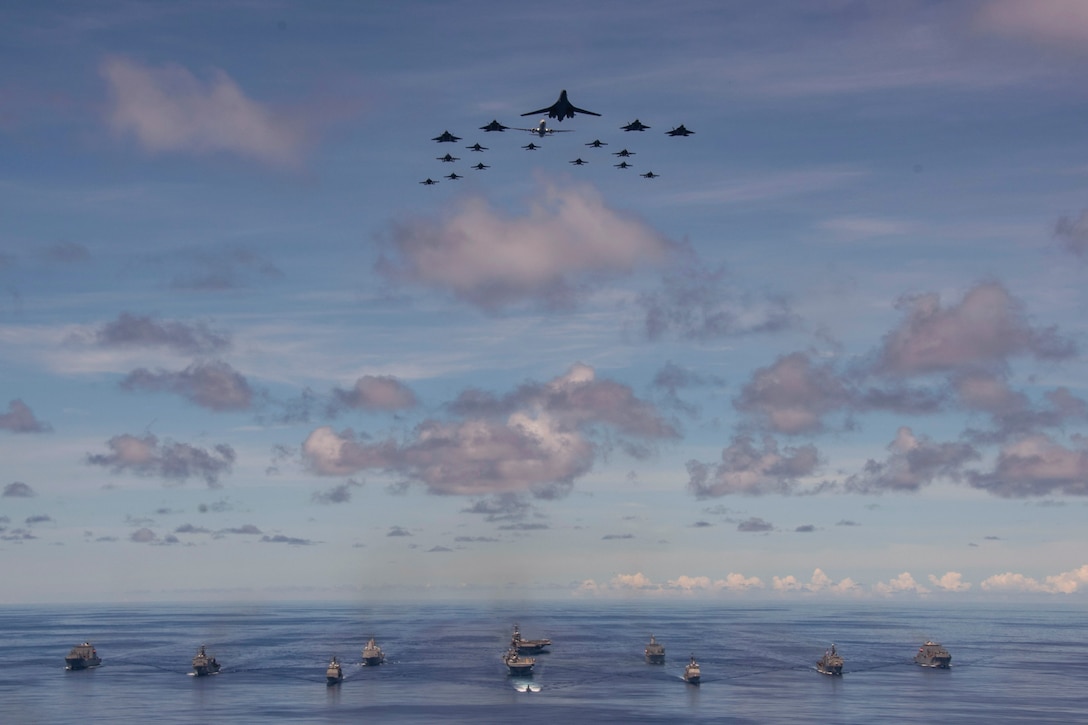 Ships sail in formation as aircraft fly above.