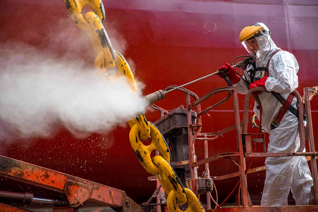 A worker in protective coveralls sprays a yellow chain in front of a red-painted ship's hull.