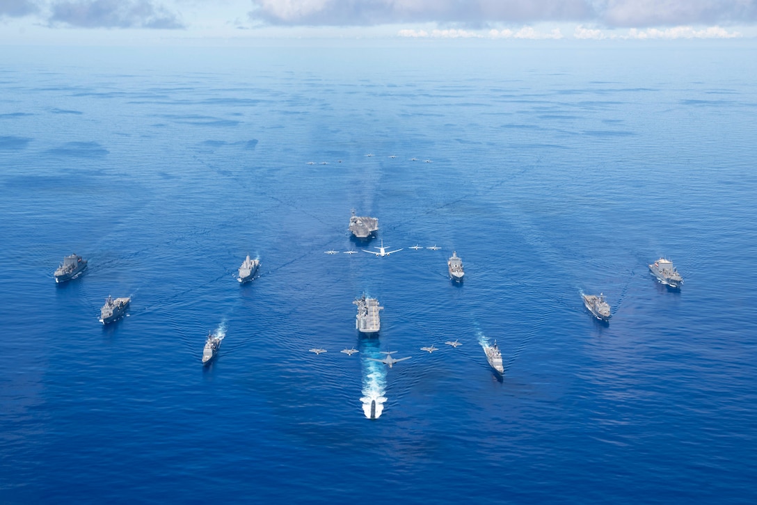 Navy ships travel in a V-type formation in vivid blue waters as Air Force and Navy aircraft fly above them.