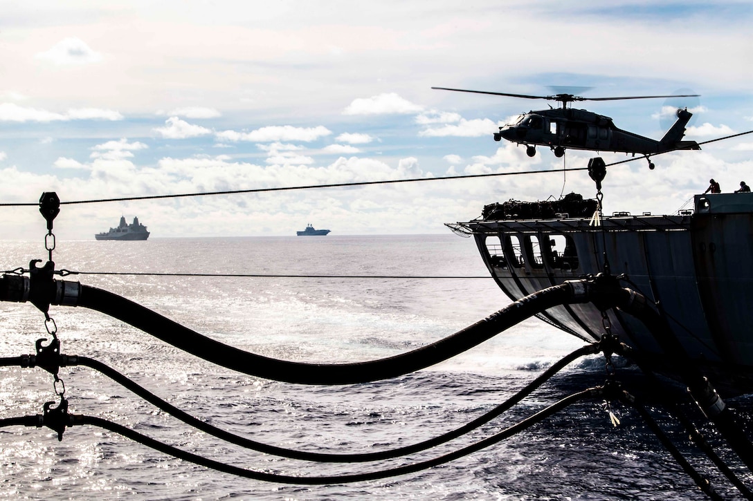 A Navy helicopter flies above a ship; two ships sail in the background.