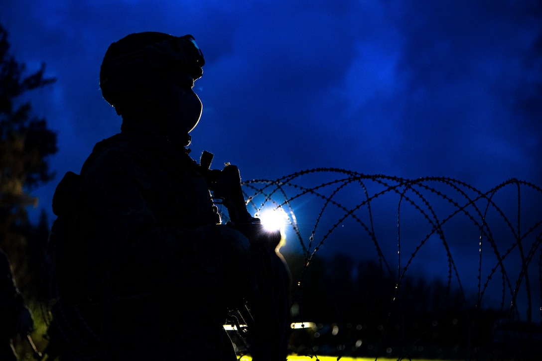 A Marine stands near some barbed wire in the darkness.