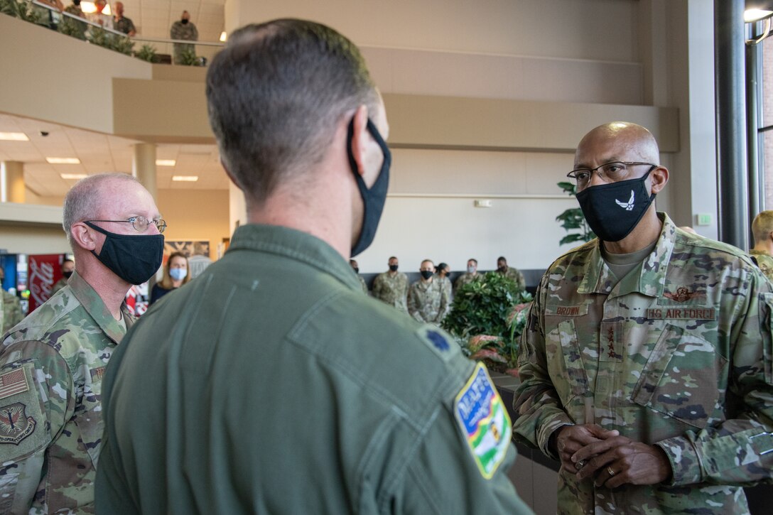 Three men wearing black masks converse in a room with a small audience in the background.