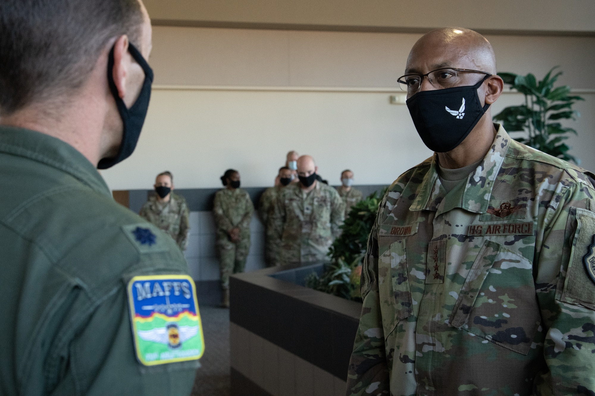 Two men converse inside a room with six onlookers in the background. Both are wearing black masks, and one is wearing an aerial firefighting patch on his flight uniform.