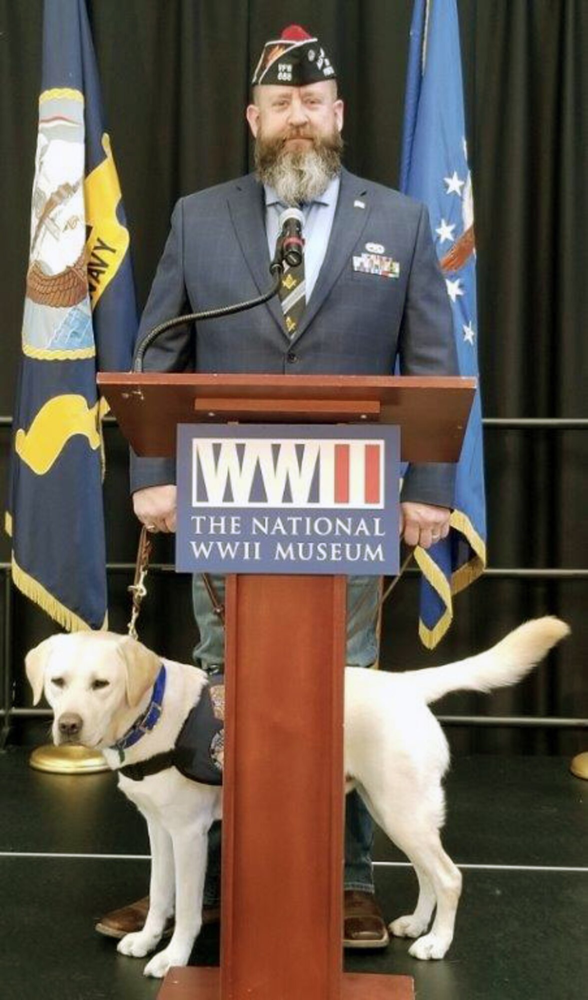 Andrew Camplen, Director of Client Services for the United States Veterans Service Dogs, provides the commencement speech while his service dog, Sebastian, stands-by at the USVSD, Dec. 2019 graduation at the National WWII Museum, New Orleans, Louisiana. The USVSD was created for and dedicated to helping veterans return to a new “Normal” by training and placing quality service dogs with veterans.