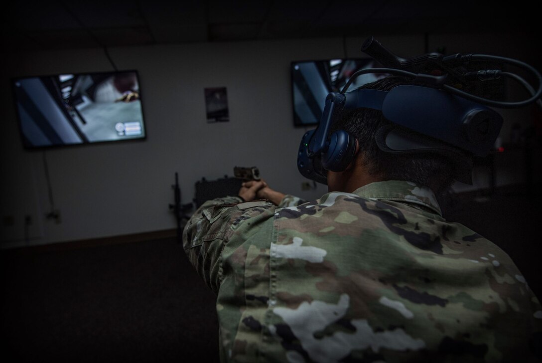 A man in a military uniform holds an orange-tipped virtual reality gun and headset in a dark room