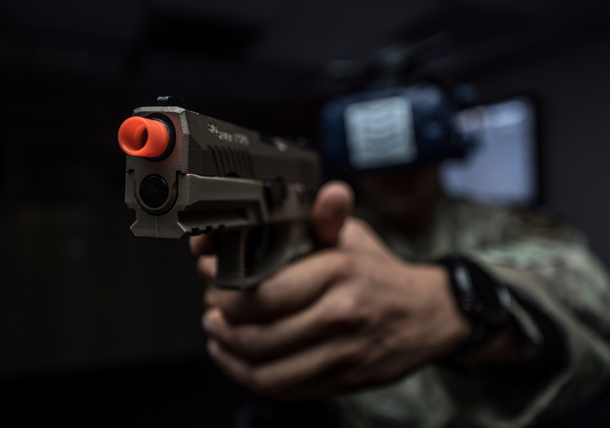 A man in a military uniform holds an orange-tipped virtual reality gun and headset in a dark room
