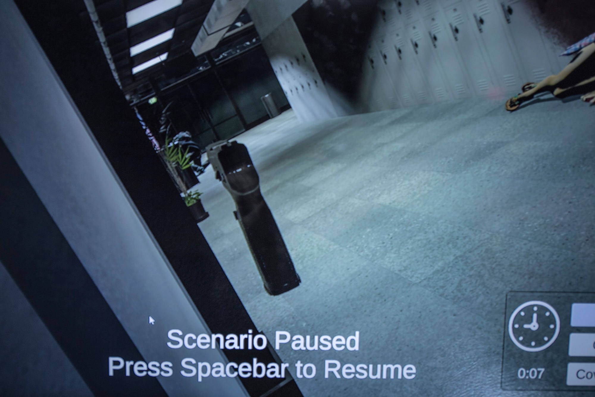 A video screen displays a first-person perspective complete with a disembodied pistol. The view is down a hallway with a crouched figure in the background.