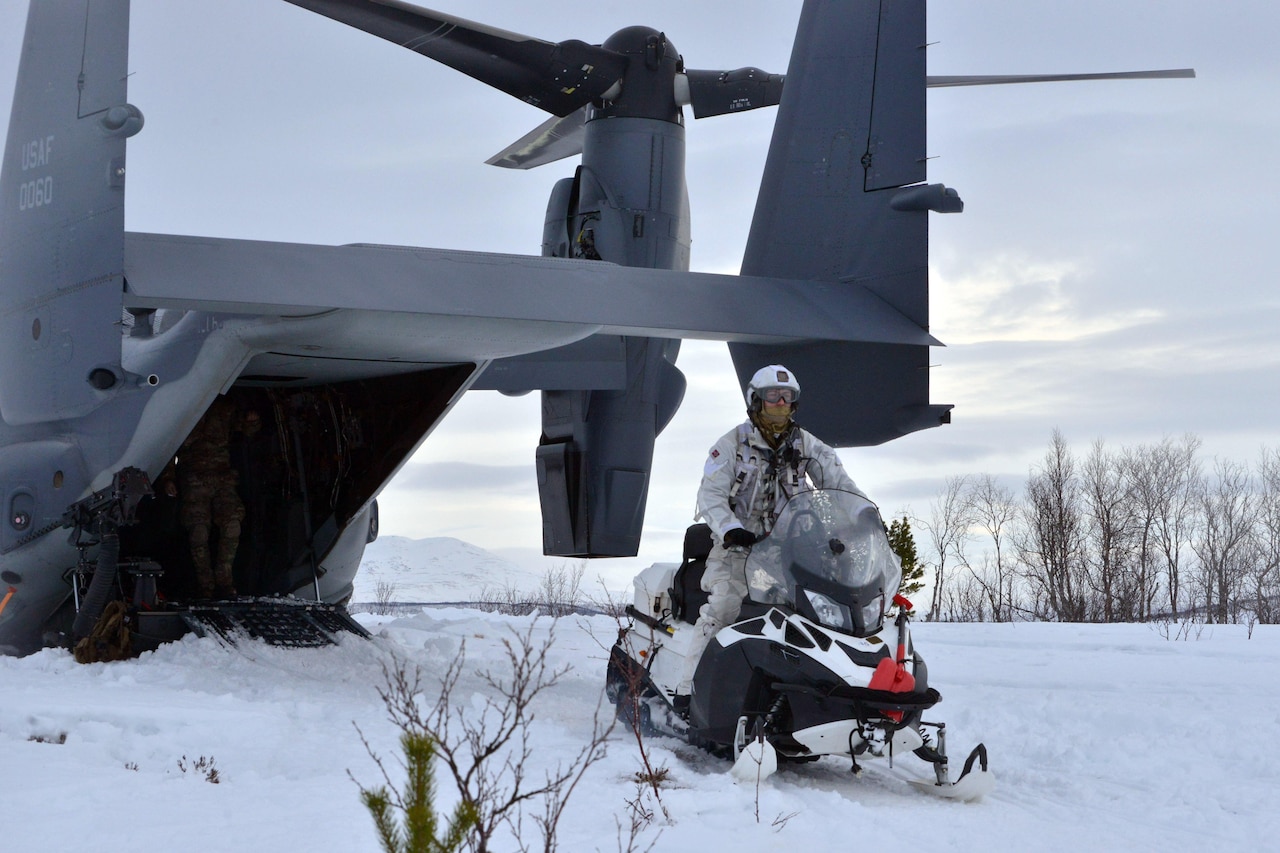 A man rides a snowmobile. A helicopter is behind him.