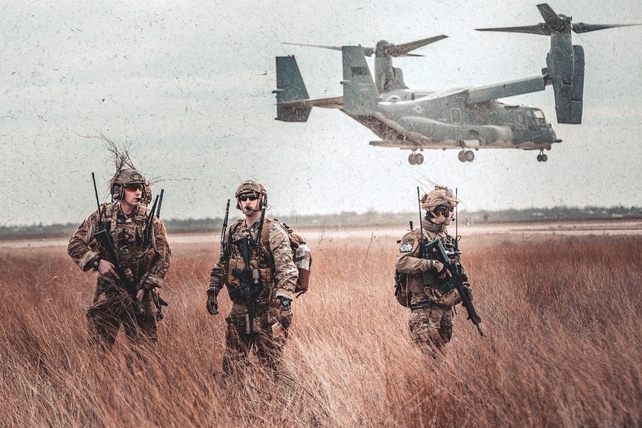Three service members with weapons walk through a field as an aircraft hovers behind them.
