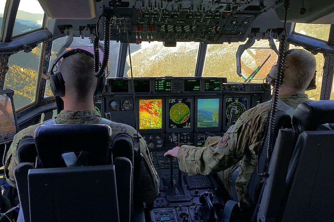 Inside a cockpit, two pilots view the control panel as they fly over rugged terrain.