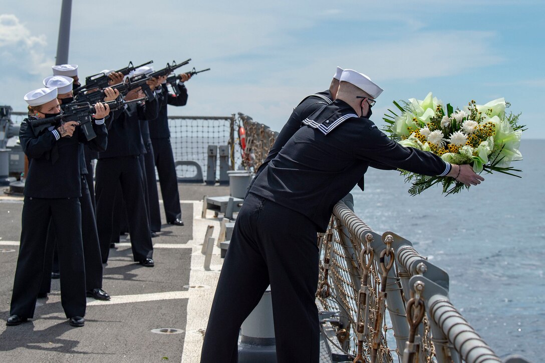 Sailors prepare to drop a wreath in the water while others shoot a volley into the air.