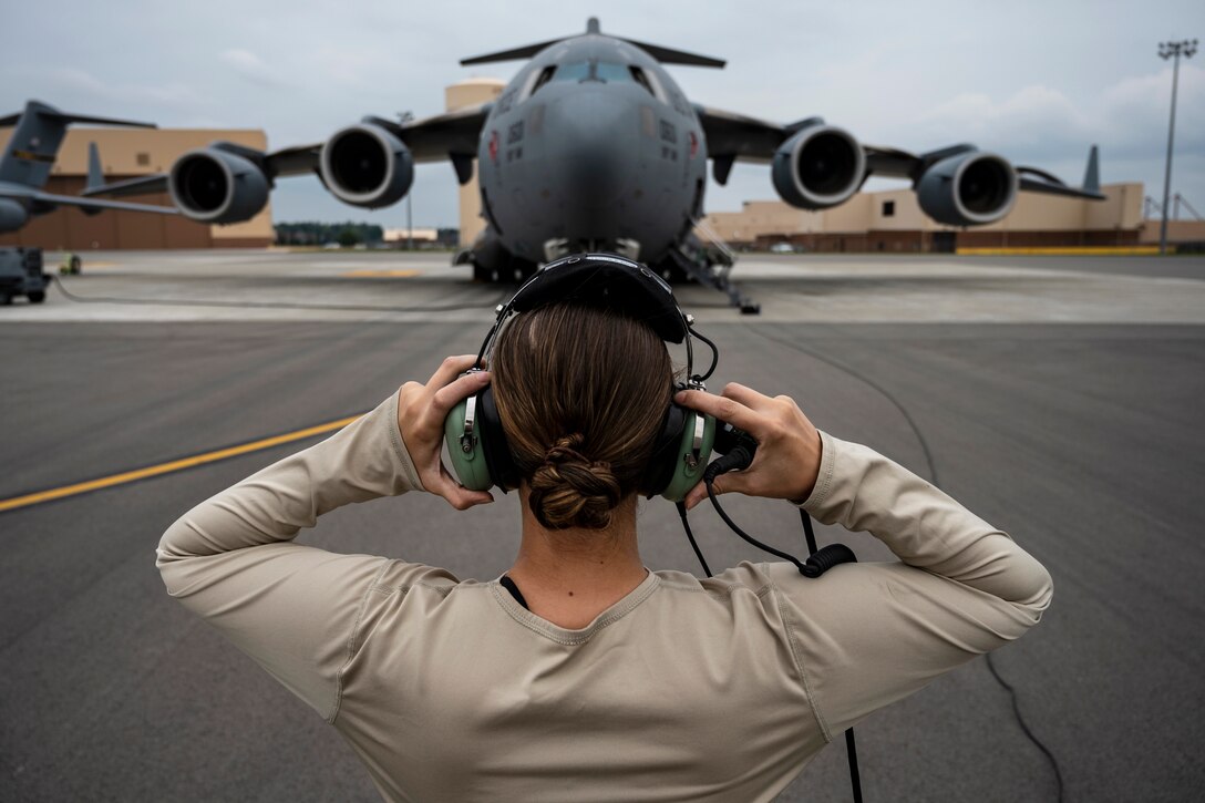An airman holds her headset while facing a large military aircraft.