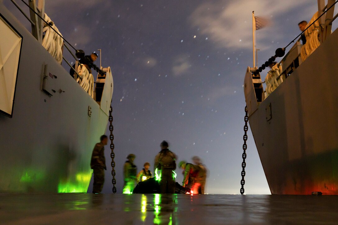 Marines load a landing craft in between two ships illuminated by green light.