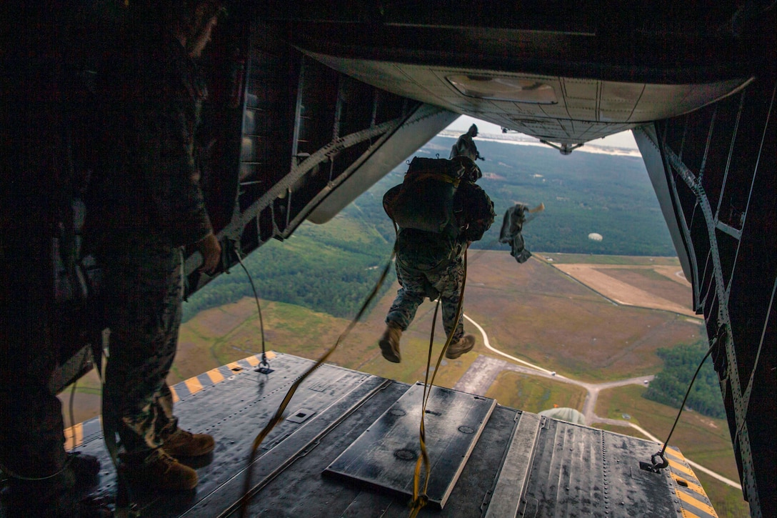 A Marine standing inside an aircraft watches another jump while wearing a parachute.