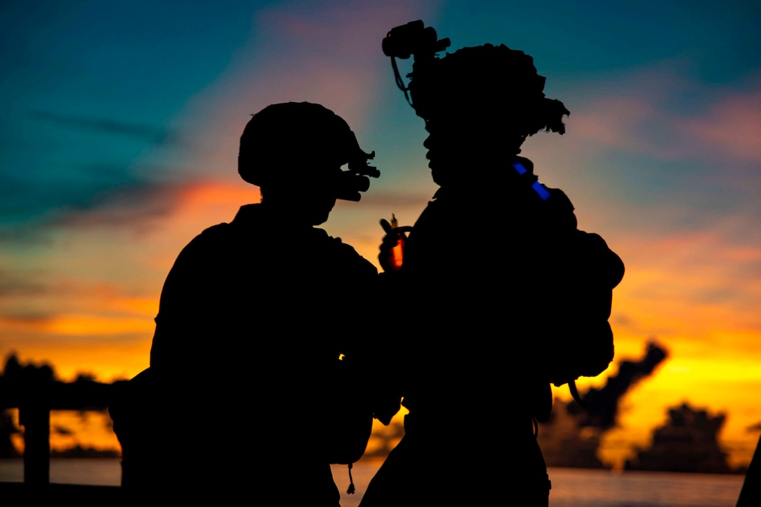 Two Marines stand together in the dark as the sun shines behind.