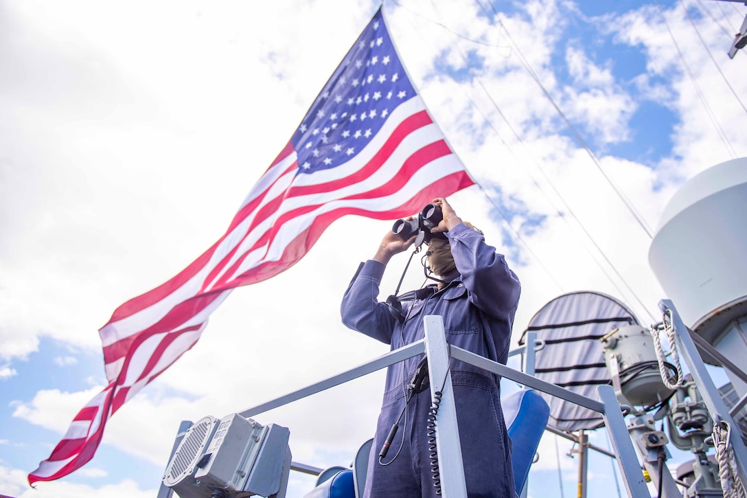 A sailor looks through binoculars as an American flag flies above.