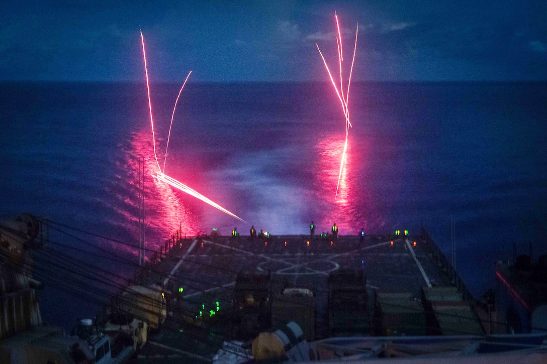 Marines fire a machine gun from a ship at night resulting in red flares across a dark ocean.