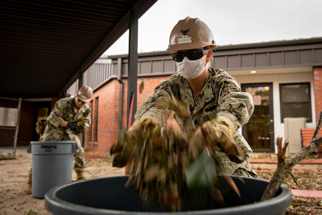 A Navy Seabee wearing a mask throws tree debris in a trash can.