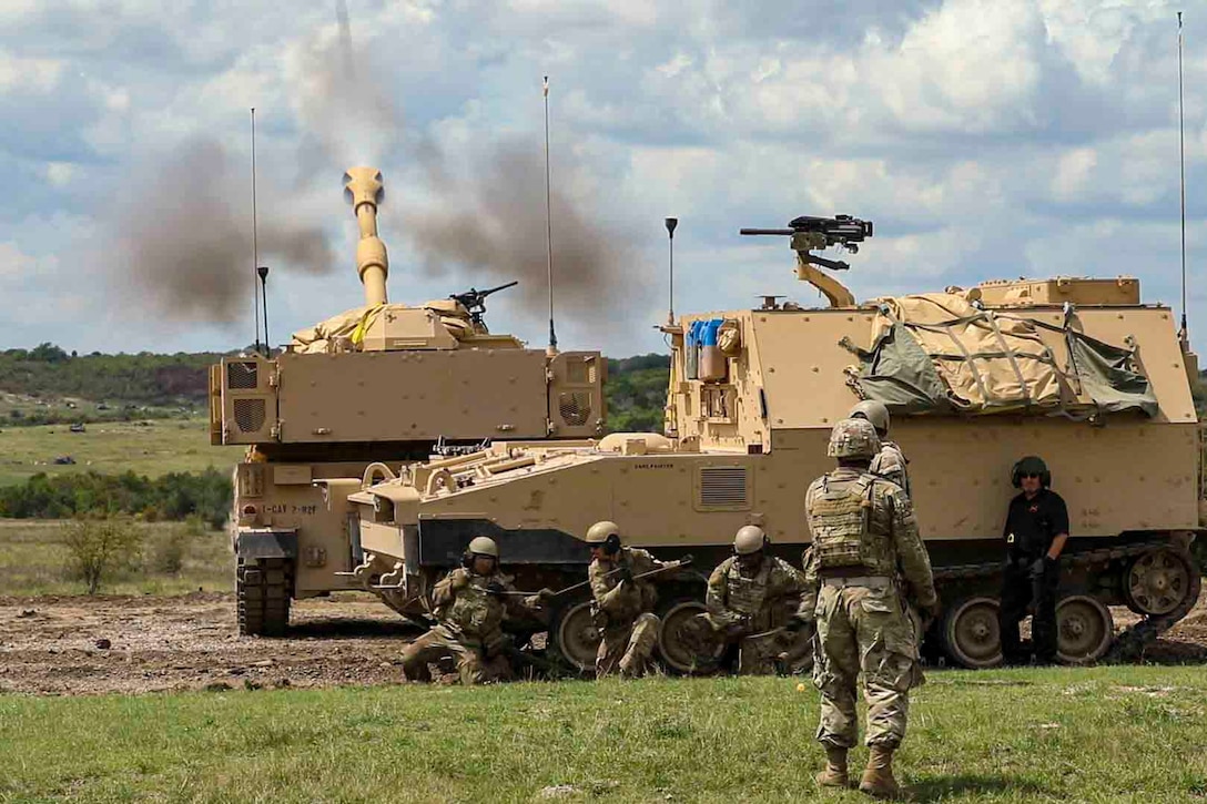 Soldiers stand behind two military vehicles as one fires artillery rounds.