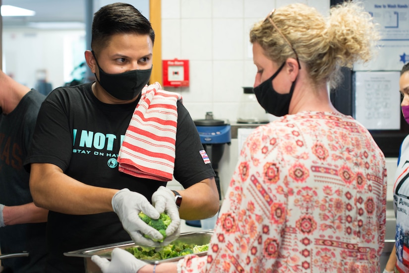 An airman wearing a face mask grabs a handful of broccoli.