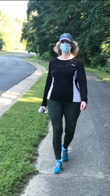 Woman wearing cap and mask walks along a sidewalk.