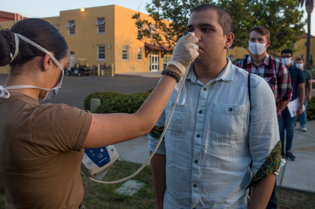 A service member wearing a face mask and gloves takes the temperature of a new recruit.
