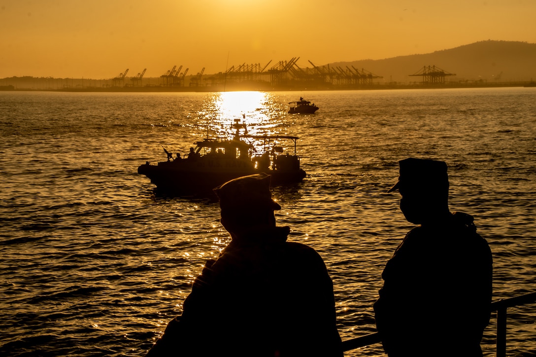 Two sailors look out at boats on the water at sunset.